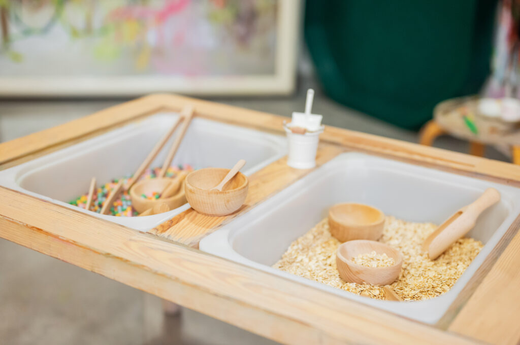 Aktywność sensoryczna Montessori w Żyrafim Domku – przesypywanie naturalnych materiałów, czerpaki i miseczki.